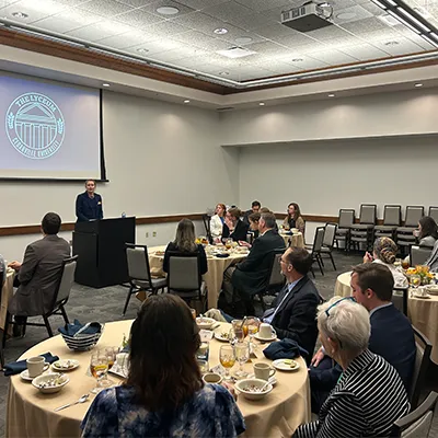 Alt text: Attendees seated at round tables listen to a speaker at a podium during a formal event in a conference room. A projection screen behind the speaker displays the Cedarville University Lyceum logo.
