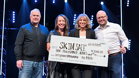 Alex Simons smiling with judges onstage holding a large check for Skin Safe.