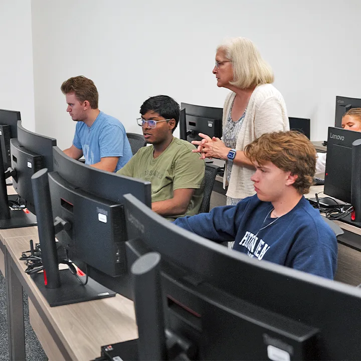 Three students working on desktop computers while professor looks on.
