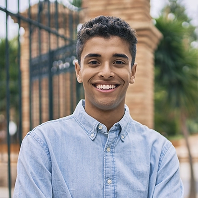 Smiling student in a light denim shirt standing outdoors near an iron fence and brick structure