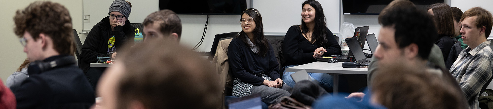 Several students seated at computers smile as they receive instructions.