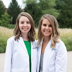 Two female pharmacy students in white coats smiling outdoors on Cedarville University’s campus