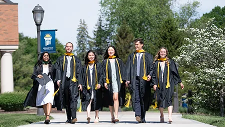 Six Cedarville University graduates in caps and gowns walk together joyfully near the campus chapel.