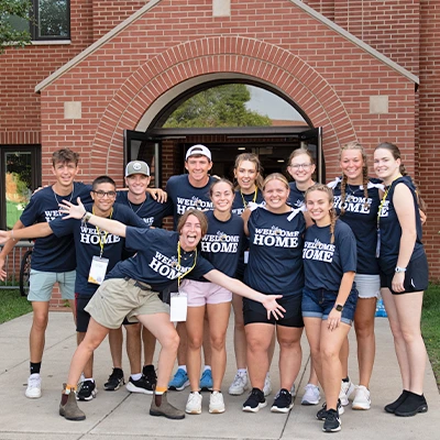 Group of 12 students wearing Welcome Home shirts smiling in front of residence hall.